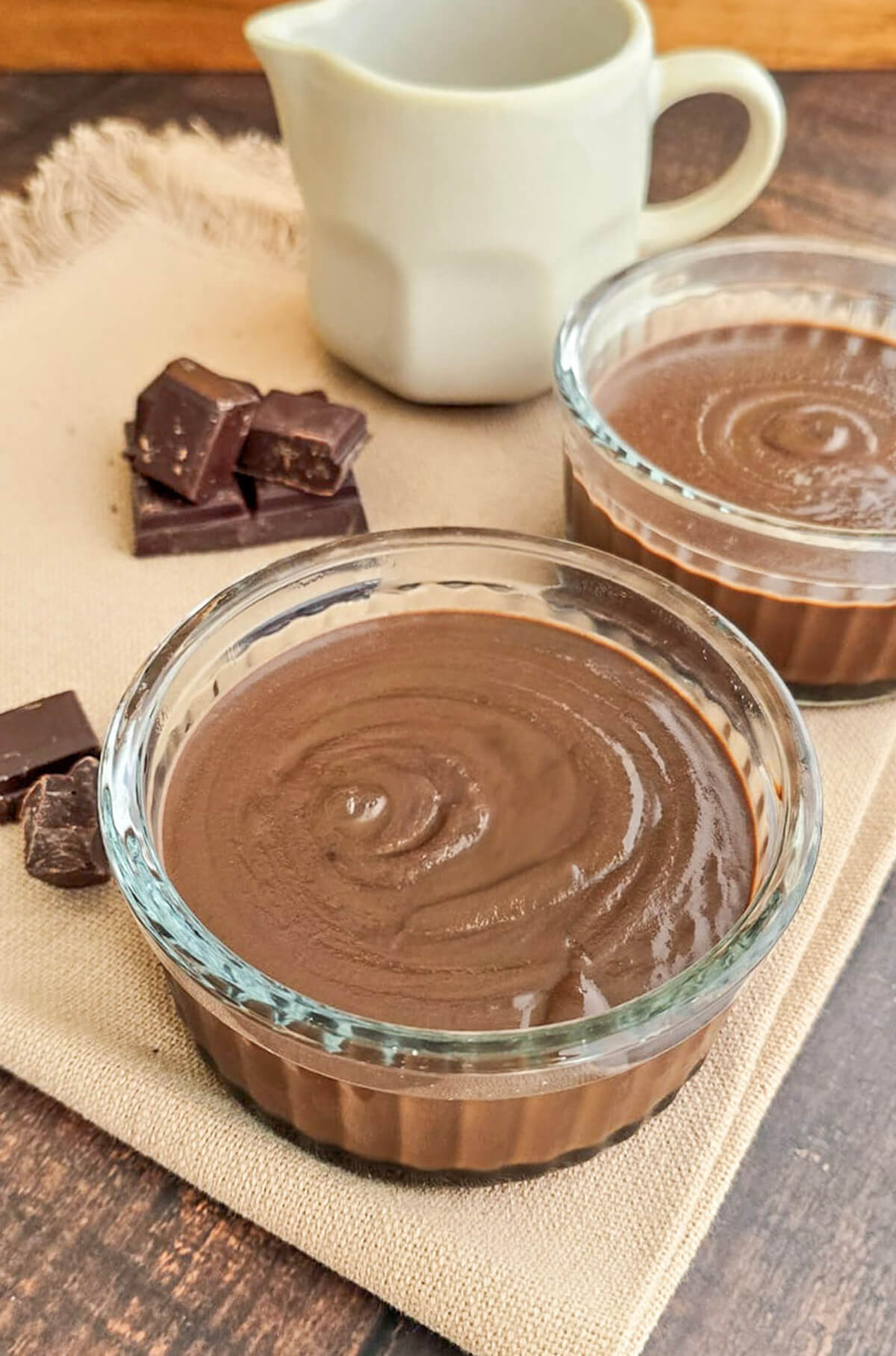 Deux ramequins en verre remplis de crème dessert onctueuse au chocolat et lait concentré, disposés sur une serviette en lin avec des carrés de chocolat noir et un pichet en céramique en arrière-plan.