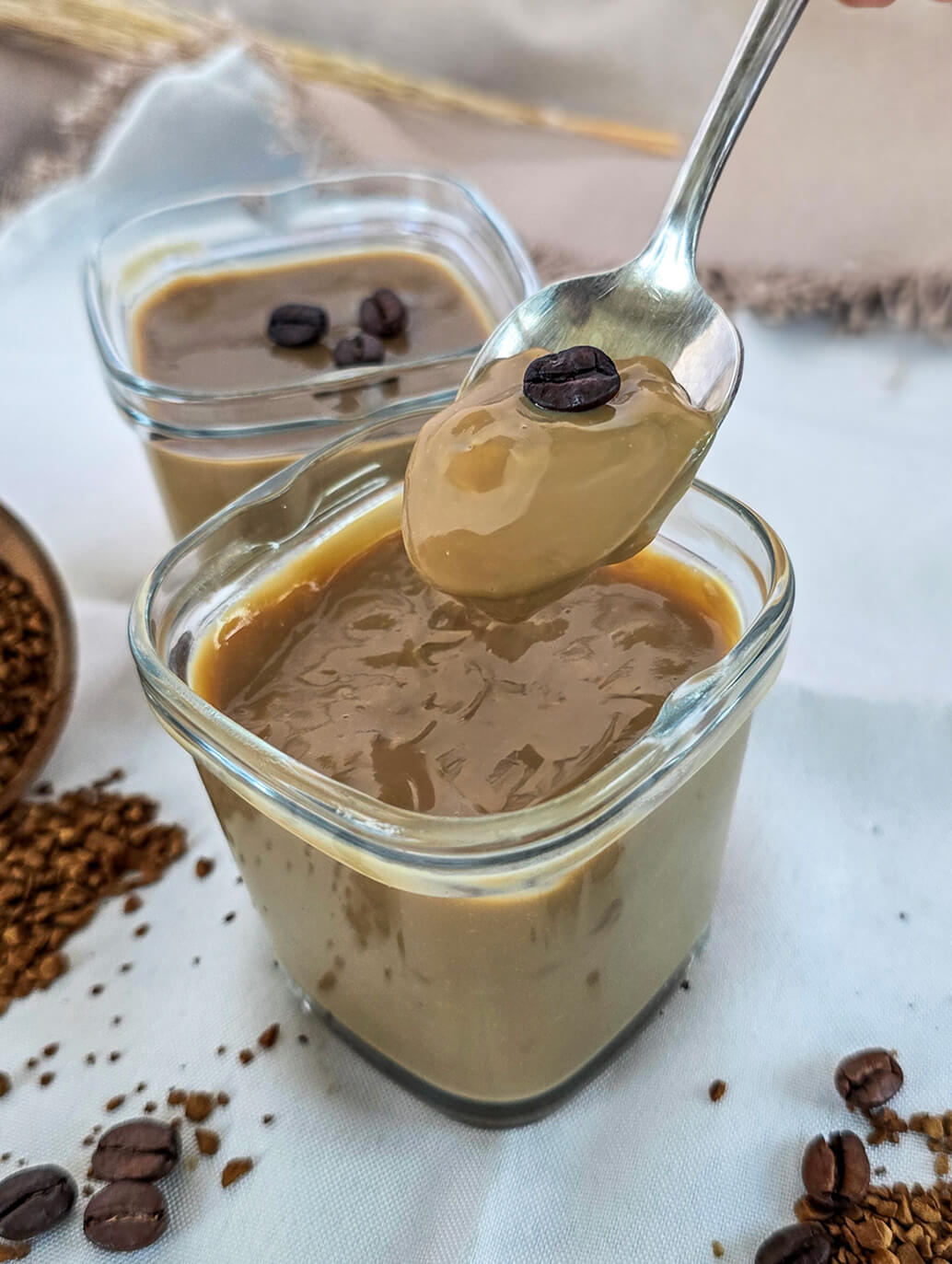 Une cuillère plongée dans une crème dessert caramel, grains de café et café soluble en poudre sur les cotés.