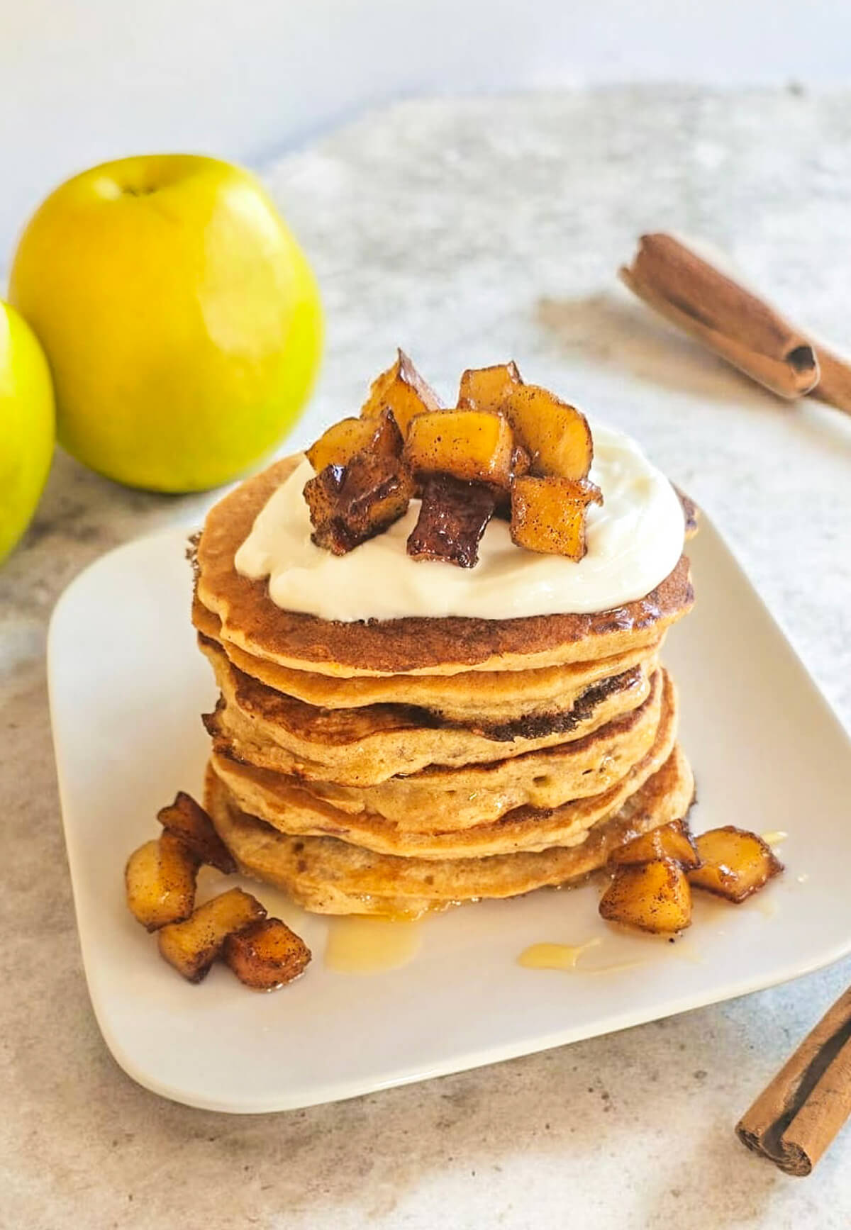 Une pile gourmande de pancakes aux pommes bien épais et dorés, surmontée de yaourt onctueux et de dés de pommes caramélisées à la cannelle. Deux pommes vertes et un bâton de cannelle sont visibles en arrière-plan sur un plan de travail clair.