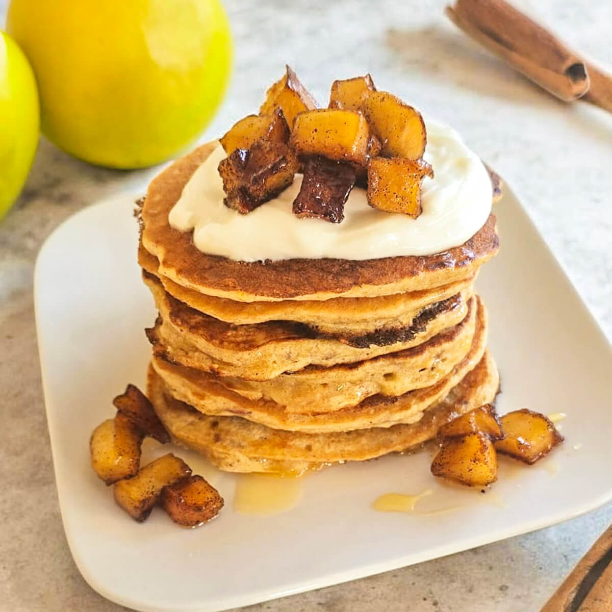 Une pile gourmande de pancakes aux pommes bien épais et dorés, surmontée de yaourt onctueux et de dés de pommes caramélisées à la cannelle. Deux pommes vertes et un bâton de cannelle sont visibles en arrière-plan sur un plan de travail clair.