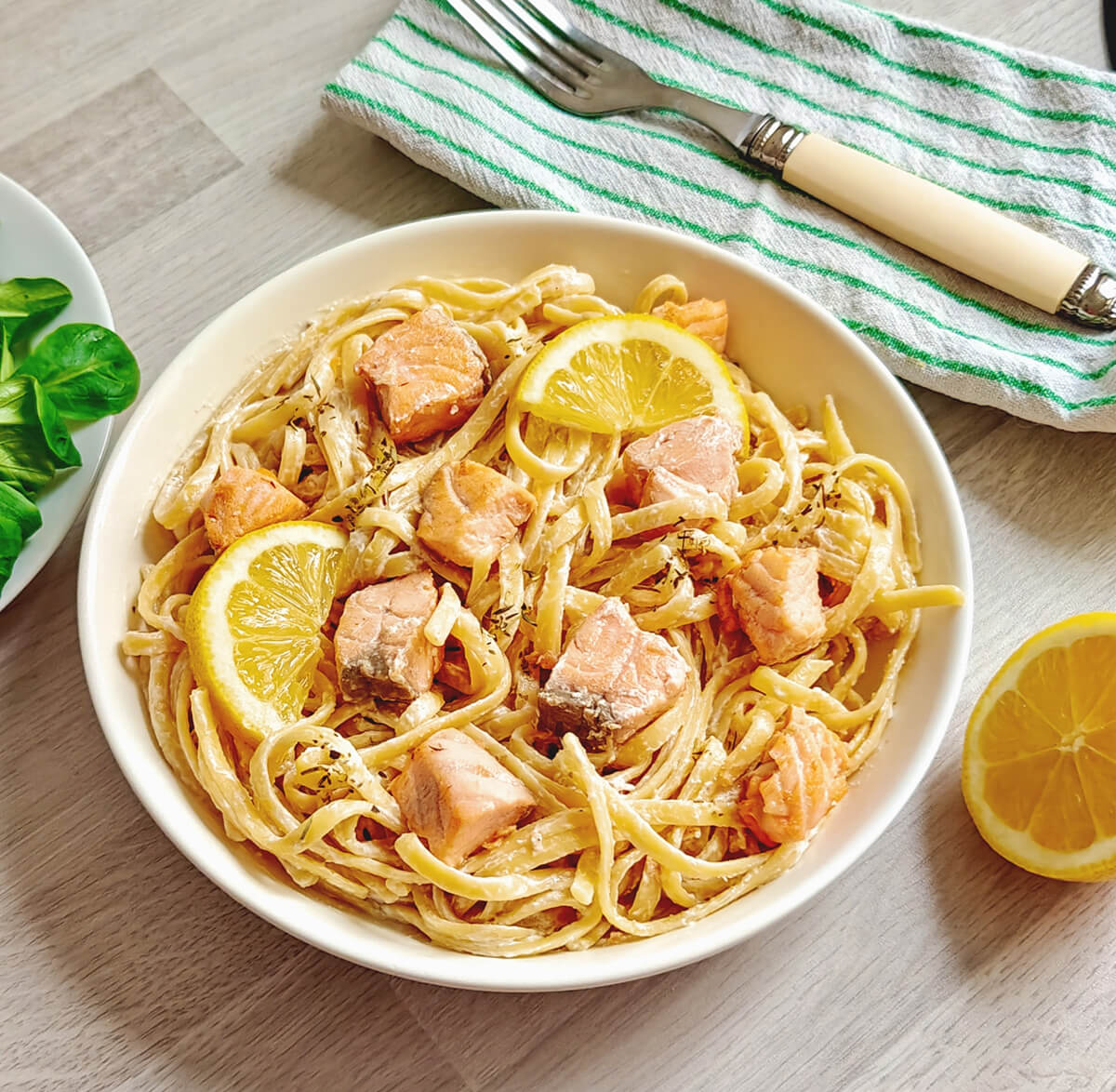 Gros plan sur une assiette de tagliatelles au saumon, sauce blanche aérienne au skyr et citron, parsemée d'herbes fraîches.