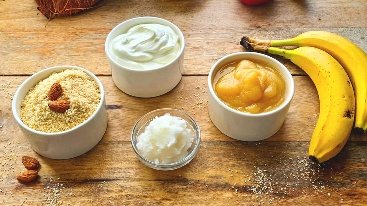 Cinq ingrédients sains pour remplacer le beurre en pâtisserie présentés dans des bols sur une table en bois : poudre d'amande, yaourt, huile de coco, compote de pommes et bananes mûres.