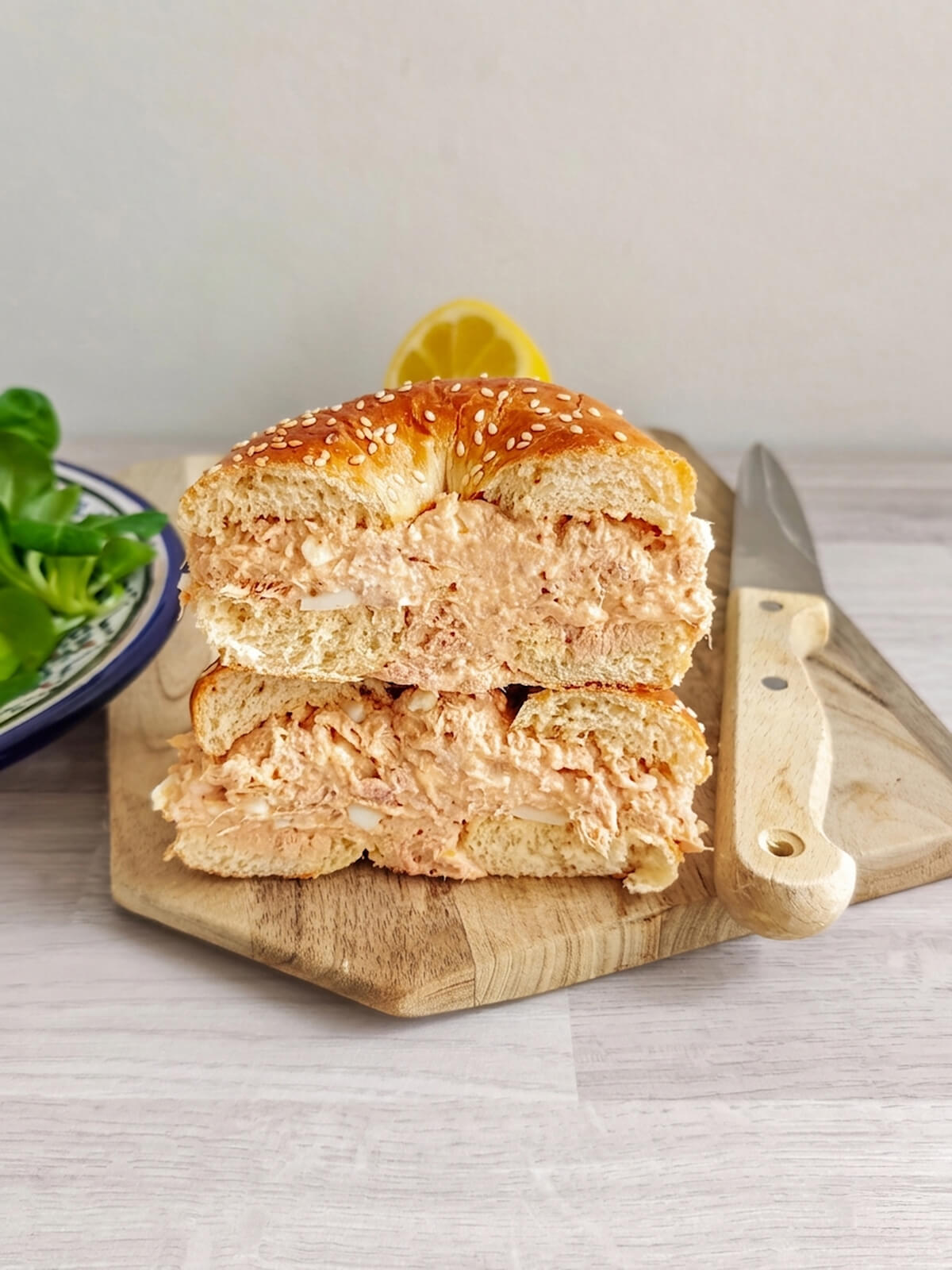 Photo d'un bagel au thon et au sésame, coupé en deux et superposé pour révéler une généreuse couche de salade de thon crémeuse. Les deux moitiés sont posées sur une planche en bois, avec un couteau à droite, un demi-citron et une assiette de mâche en arrière-plan.