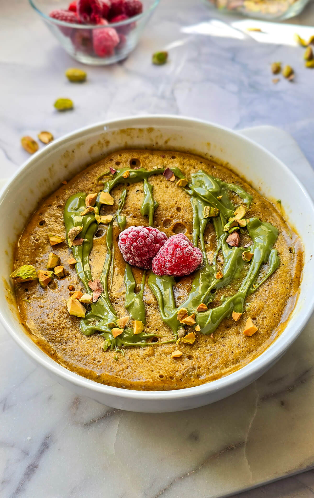 Vue rapprochée d'un bowl cake à la pistache, garni de deux framboises surgelées au centre et de pistaches concassées, avec un filet de crème de pistache. Arrière-plan flou de framboises et pistaches sur un plan de travail en marbre.