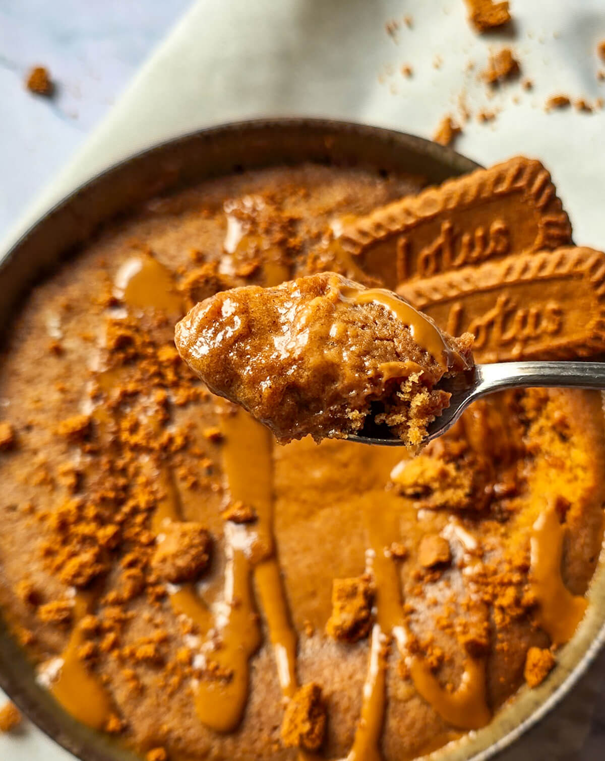 Gros plan sur une cuillère remplie de bowlcake moelleux au spéculoos, brillant de nappage fondant. En arrière-plan, le bol et des biscuits Lotus entiers.