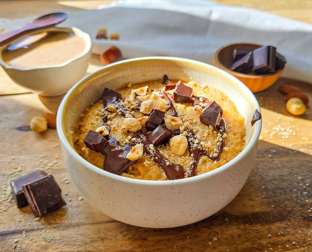 Un mug cake choco-noisettes dans un bol en céramique moucheté, nappé de chocolat fondu, d'éclats de chocolat noir et de noisettes entières et concassées. Le bol repose sur une planche en bois à côté de carreaux de chocolat et de noisettes éparpillés.