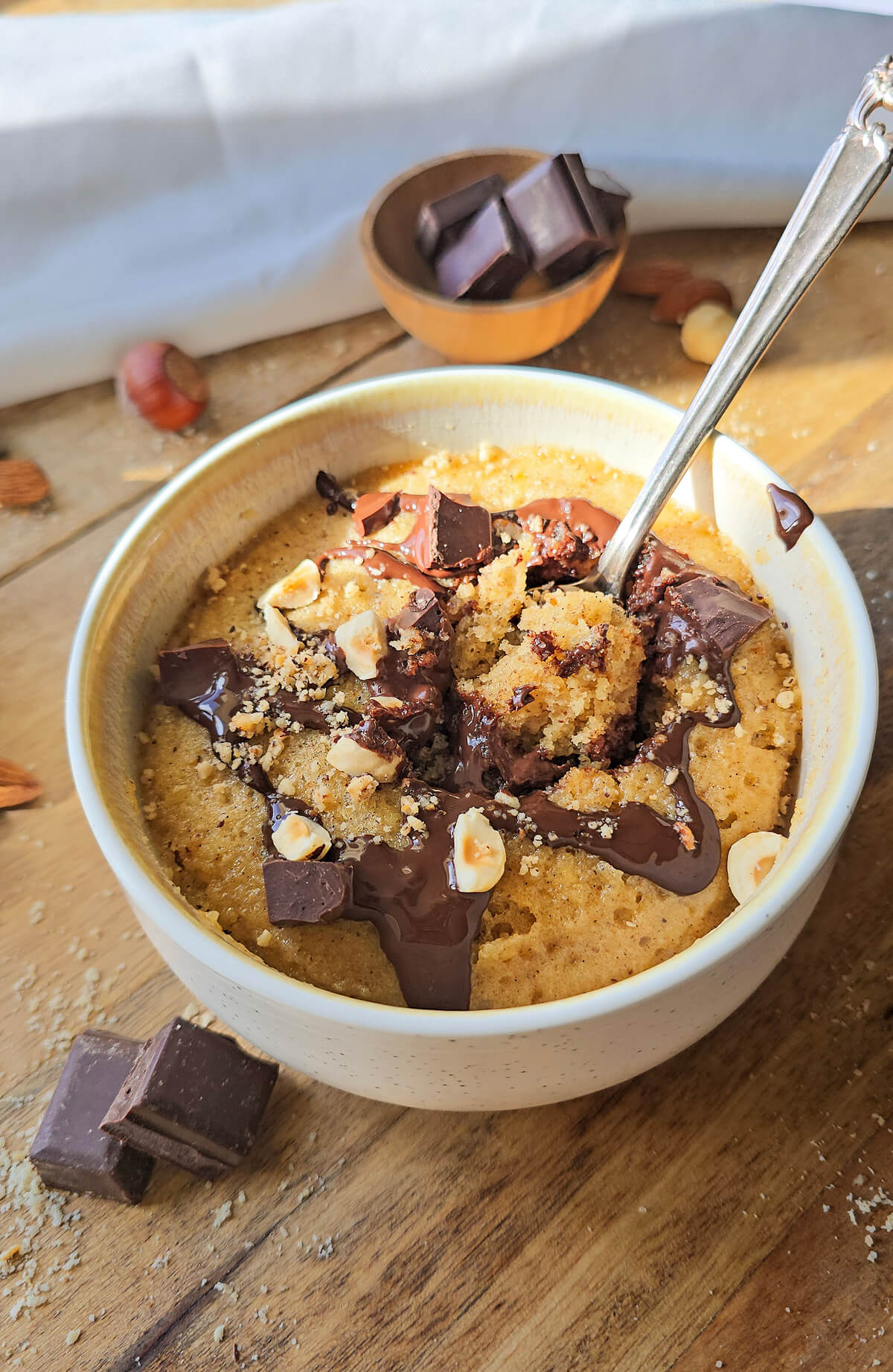 Le même mug cake choco-noisettes, cette fois avec une cuillère en argent plongeant à l'intérieur, révélant une mie moelleuse et humide. Le nappage au chocolat fondu et les toppings sont bien visibles.