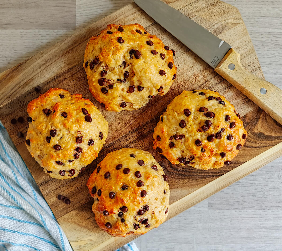 Photographie aérienne de quatre briochettes pépites de chocolat dorées et moelleuses, disposées en carré sur une planche à découper en bois rustique. Une lame de couteau est posée en diagonale sur la planche et un linge rayé bleu et blanc est visible sur le côté.