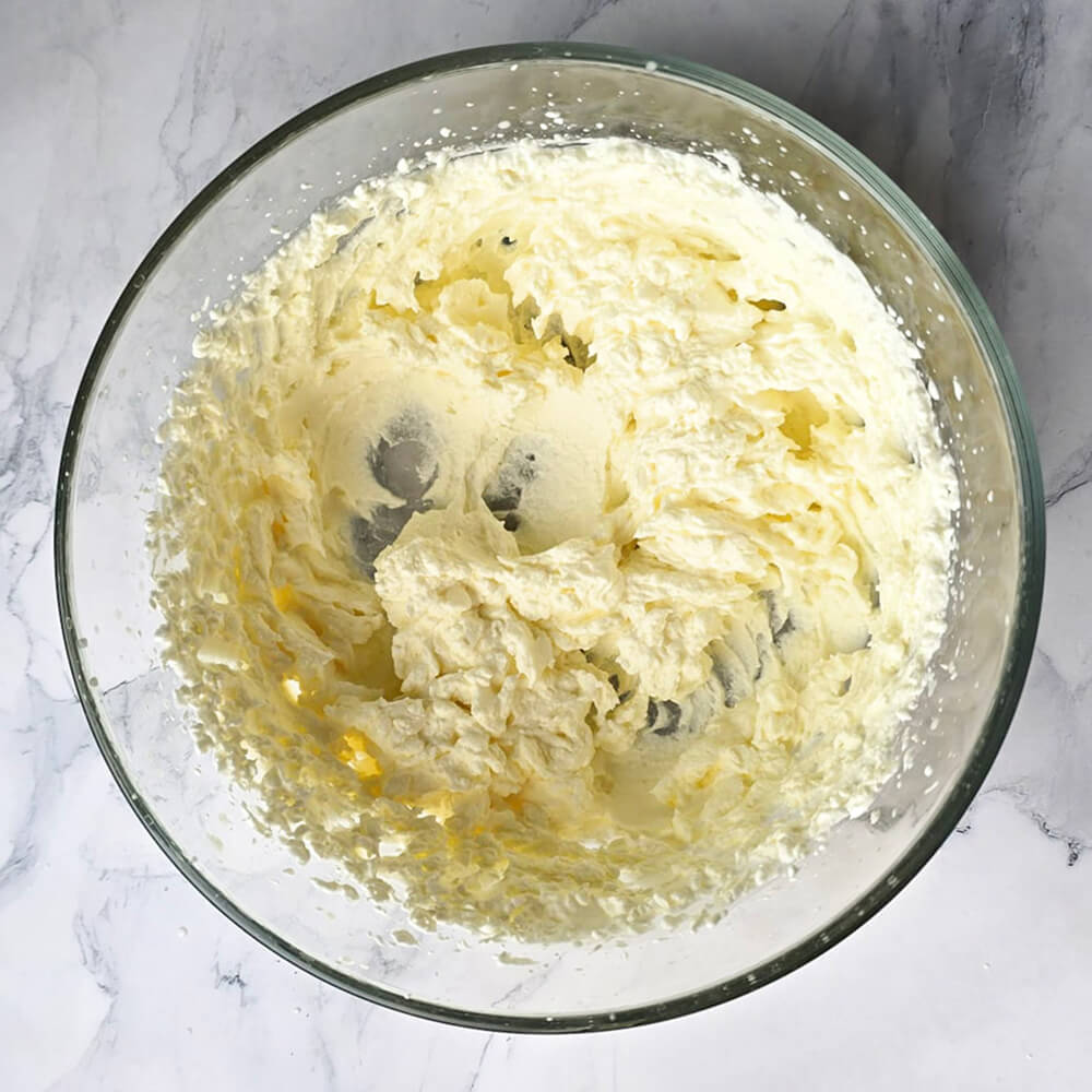 Une vue de dessus d'un bol en verre transparent contenant de la crème liquide entière qui a tranché, montrant une texture grumeleuse et jaune pâle éparpillée au fond, signe d'une chantilly ratée prête à être transformée.