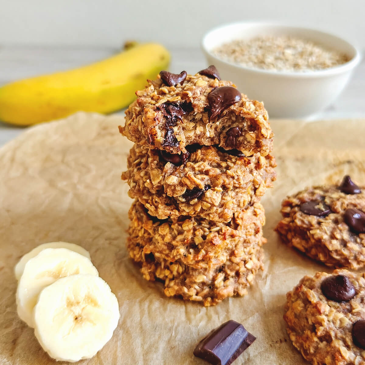 Une pile de trois cookies moelleux aux flocons d'avoine et à la banane, parsemés de pépites de chocolat fondant, présentée sur du papier cuisson avec des rondelles de banane à côté.
