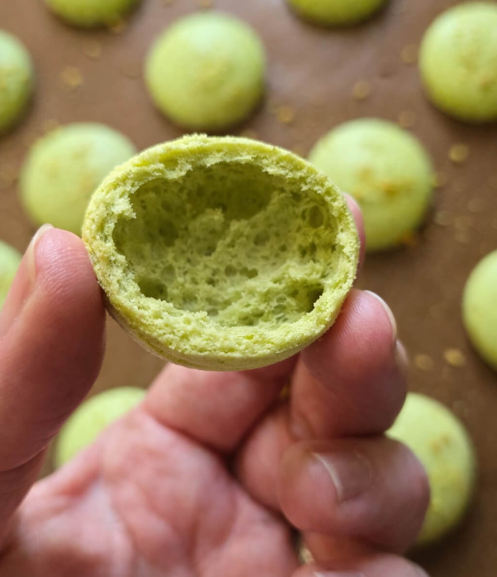 Gros plan sur une coque de macaron vert pistache coupée en deux et tenue à la main, révélant une mie moelleuse et humide à l'intérieur. En arrière-plan flou, d'autres coques de macarons cuisent sur une plaque de cuisson recouverte de papier sulfurisé