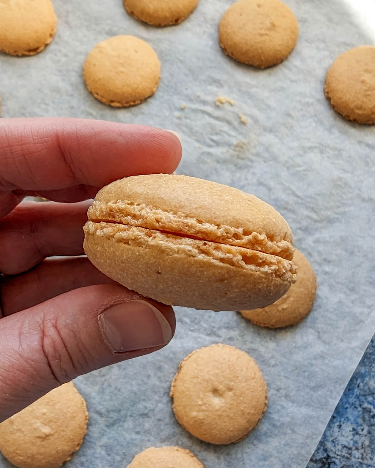 Une main tenant un macaron raté de couleur beige clair. Le macaron est un échec car le four était trop chaud. Plusieurs autres coques de macarons imparfaites sont visibles sur une plaque de cuisson recouverte de papier sulfurisé.