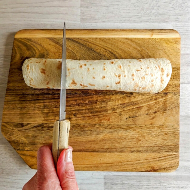 Une main tenant un couteau de cuisine en train de couper une tortilla de banane chocolat, sur une planche de bois.