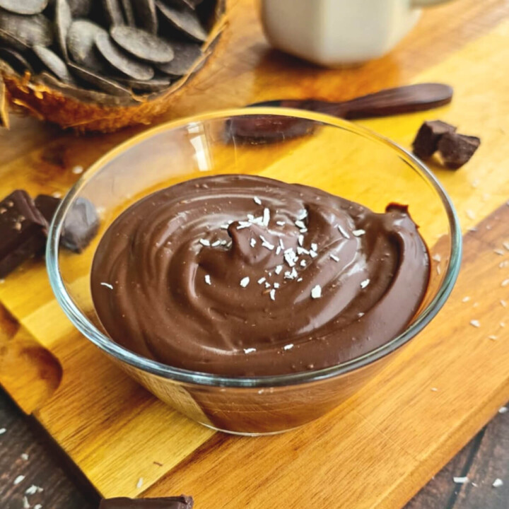 Crème dessert onctueuse au chocolat et lait de coco servie dans un bol en verre, saupoudrée de copeaux de noix de coco, présentée sur une planche en bois avec des carrés de chocolat noir et une coque de noix de coco remplie de pistoles de chocolat.