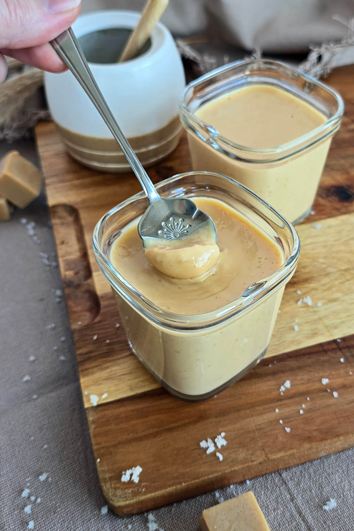 Une cuillère plongée dans une crème dessert caramel dans un petit pot de yaourtière, sur une planche de bois.