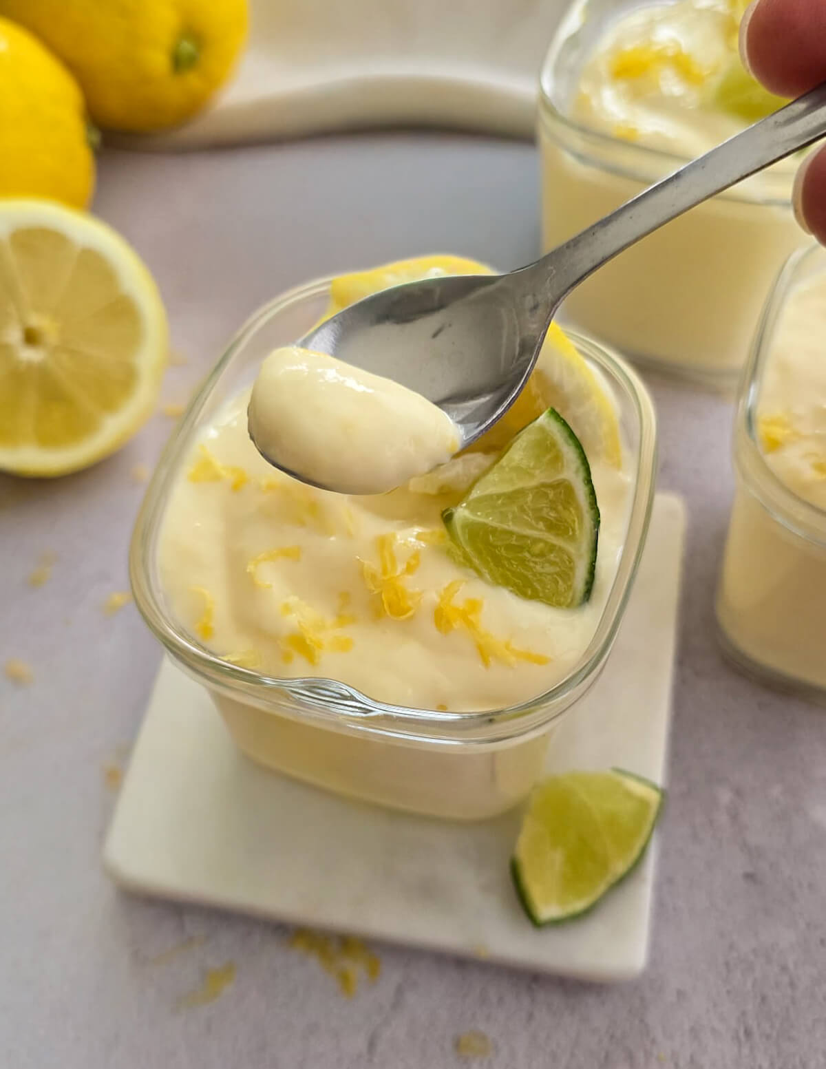 Un gros plan vertical montre une cuillère en métal prélevant une cuillerée de crème dessert au citron onctueuse d'une verrine carrée en verre. La crème est jaune pâle et parsemée de zestes de citron. Une tranche de citron vert est posée sur la crème. Deux autres verrines et des citrons entiers et coupés sont visibles en arrière-plan flou.