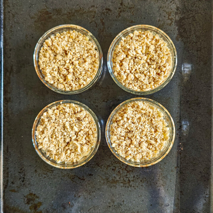 Quatre ramequins en verre garnis de pommes et recouverts de pâte au crumble disposés sur une plaque de cuisson avant d'être enfournés.