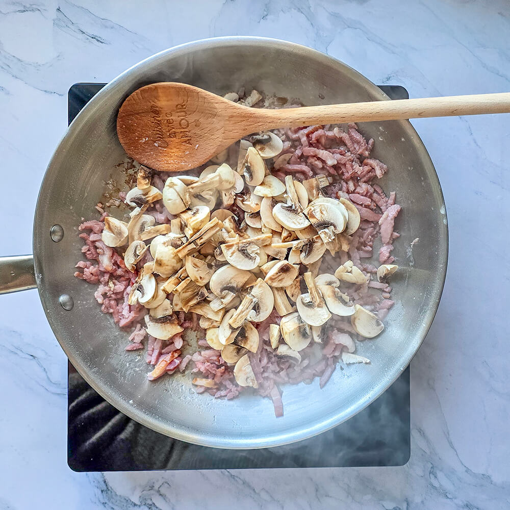 Cuisson des lardons et champignons dans une poêle en inox, avec une cuillère en bois.