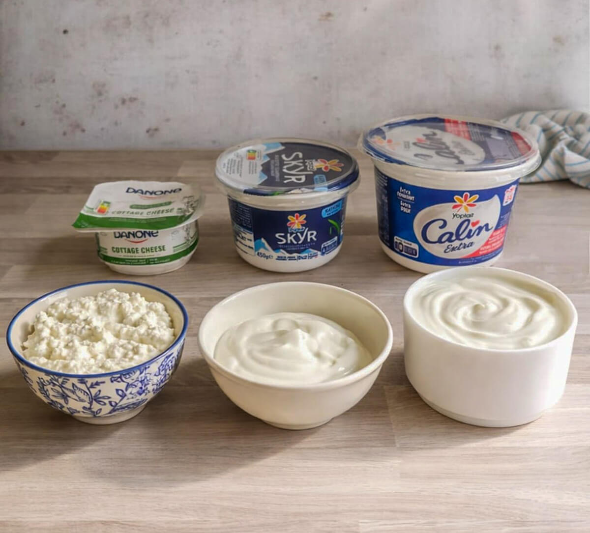 Une photo d'un plan de travail de cuisine alignant trois pots de produits laitiers de marques spécifiques (un petit Danone Cottage Cheese, un Skyr Siggi's moyen, un grand pot de Yoplait Calin Extra) et trois bols ouverts devant eux. Les bols révèlent la texture distinctive de chacun : à gauche, le cottage cheese grumeleux dans un bol bleu et blanc ; au centre, le skyr épais dans un bol beige ; à droite, le fromage blanc lisse dans un bol blanc.