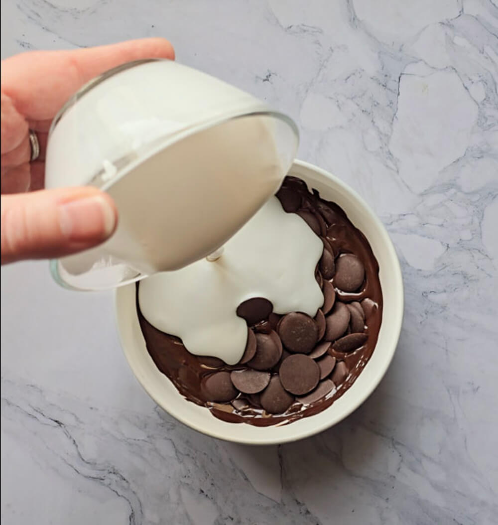 Une main verse un bol de crème de coco sur un bol de pistoles de chocolat noir, qui fondent sur un plan de travail en marbre.