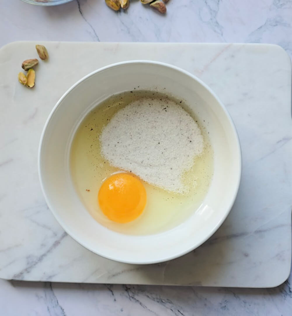 Vue de dessus d'un bol blanc sur une planche de marbre, contenant un œuf entier cru et une portion de sucre vanillé ou semoule. Arrière-plan flou de marbre et de pistaches décoratives. Première étape de la préparation.