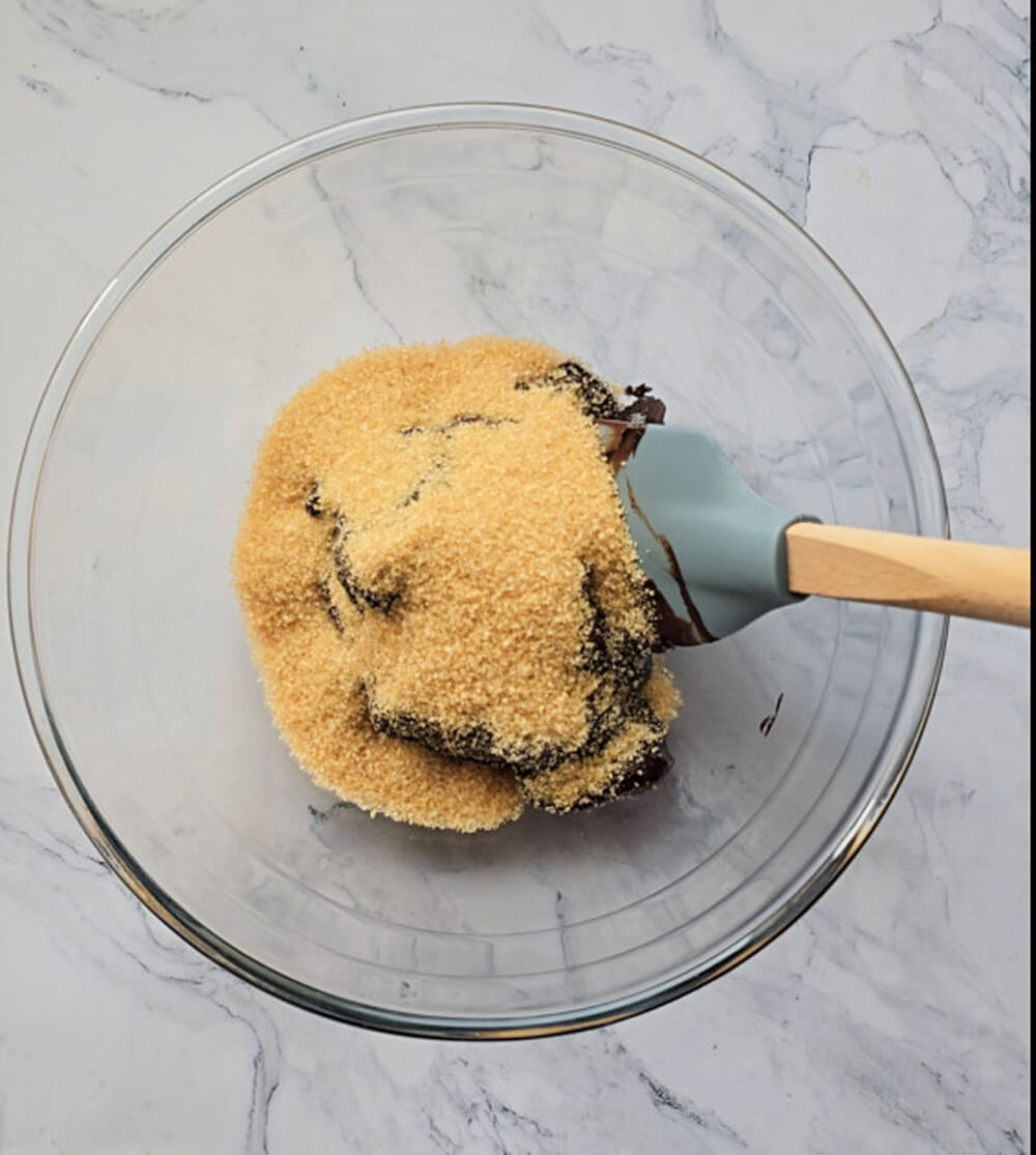 Un bol en verre rempli d'un mélange de chocolat noir fondu et de sucre roux, avec une spatule bleu clair à manche en bois.