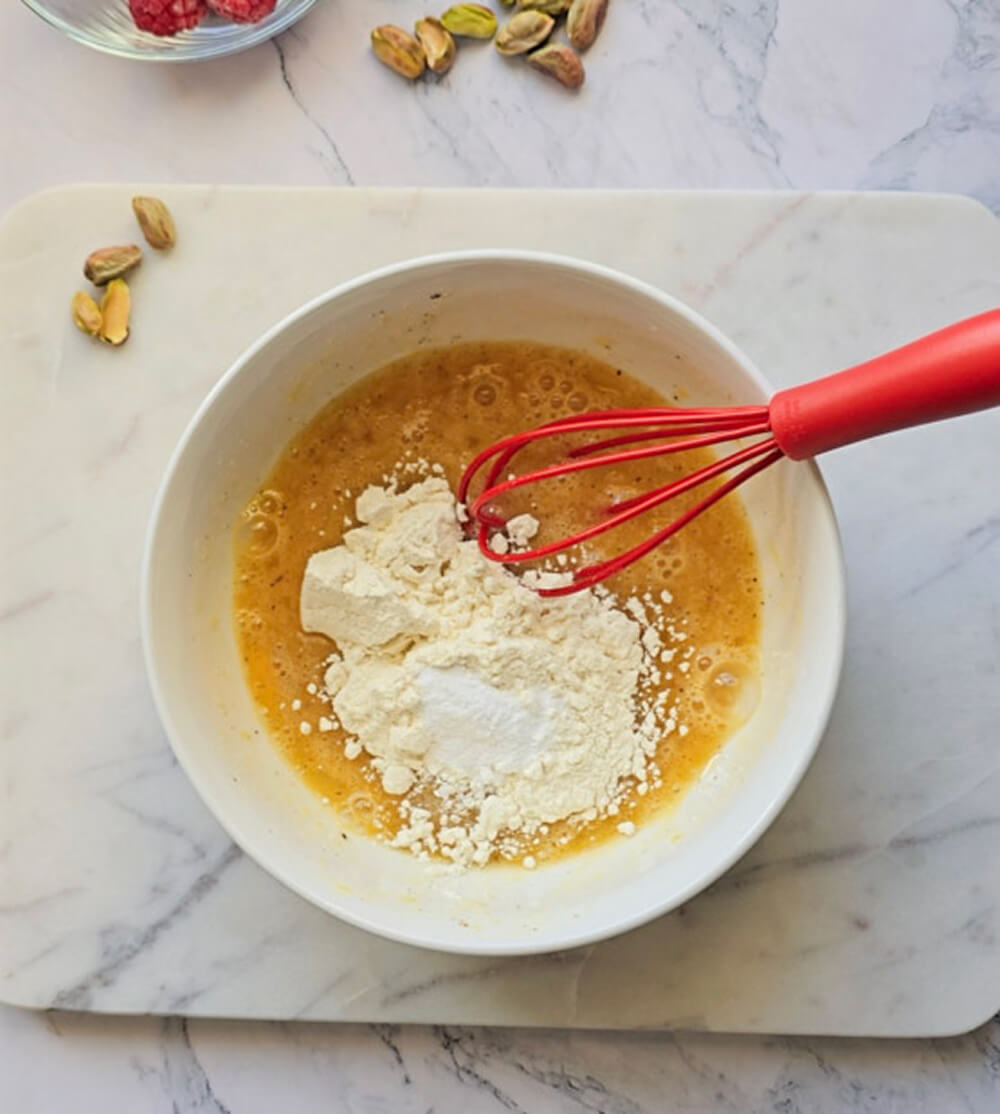 Gros plan de dessus sur le bol blanc. La pâte de base jaune est prête. Une grosse noisette de pâte de pistache verte et onctueuse est ajoutée au centre, avec un fouet rouge prêt à la mélanger. Arrière-plan de marbre avec des pistaches.