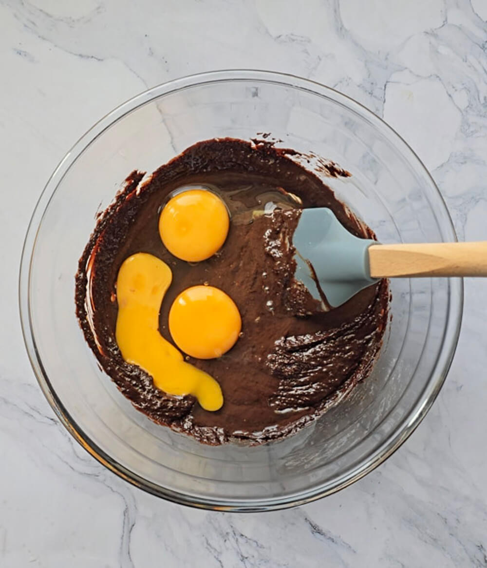 Deux jaunes d'œufs crus posés sur une pâte au chocolat noir dans un bol en verre, avec une spatule bleu clair à manche en bois.