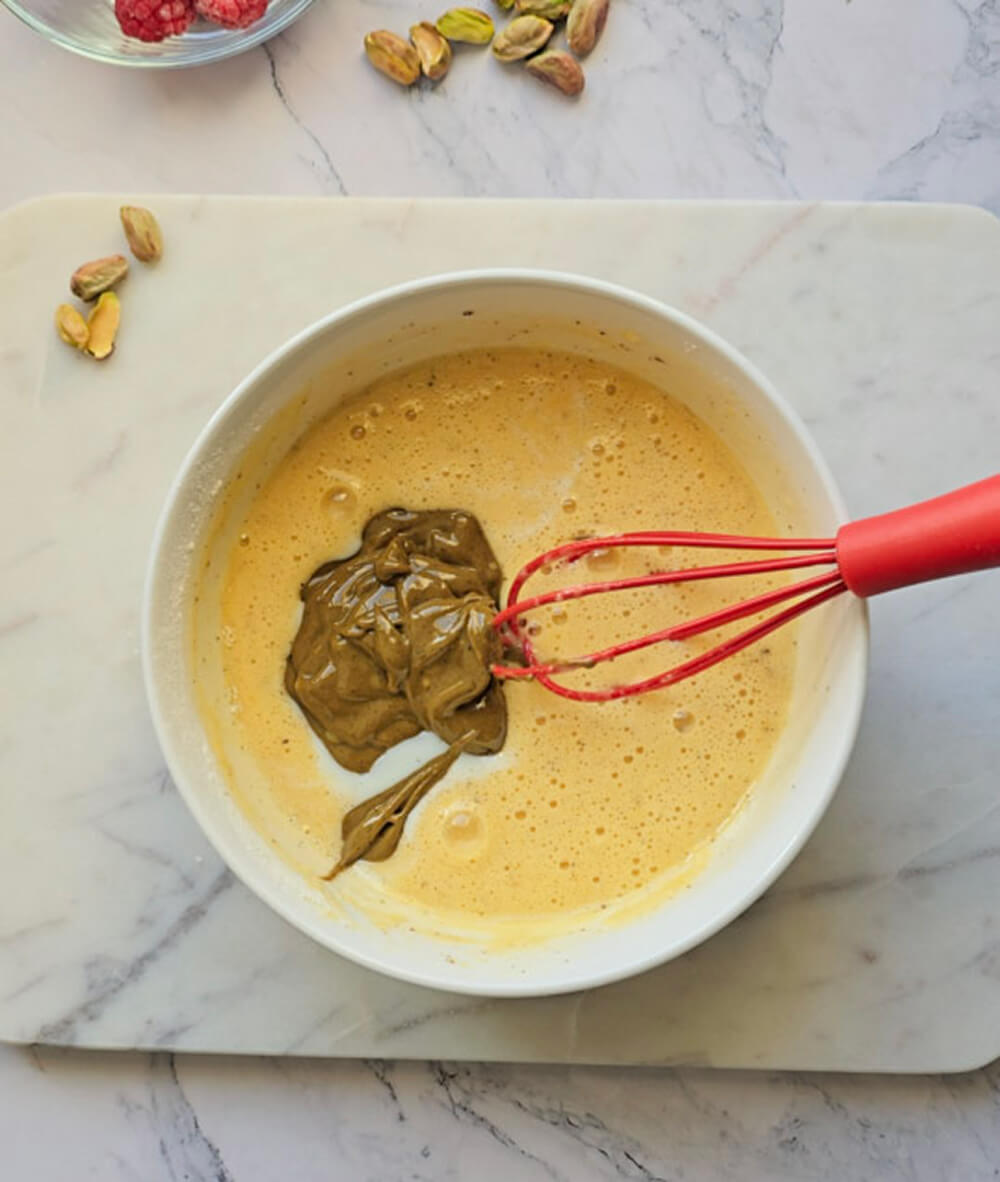 Vue de dessus du bol blanc. Le mélange est devenu jaune clair. Une pile de farine blanche tamisée et une petite pincée de levure sont ajoutées. Le fouet rouge est positionné pour incorporer les ingrédients secs. Arrière-plan de marbre avec pistaches.