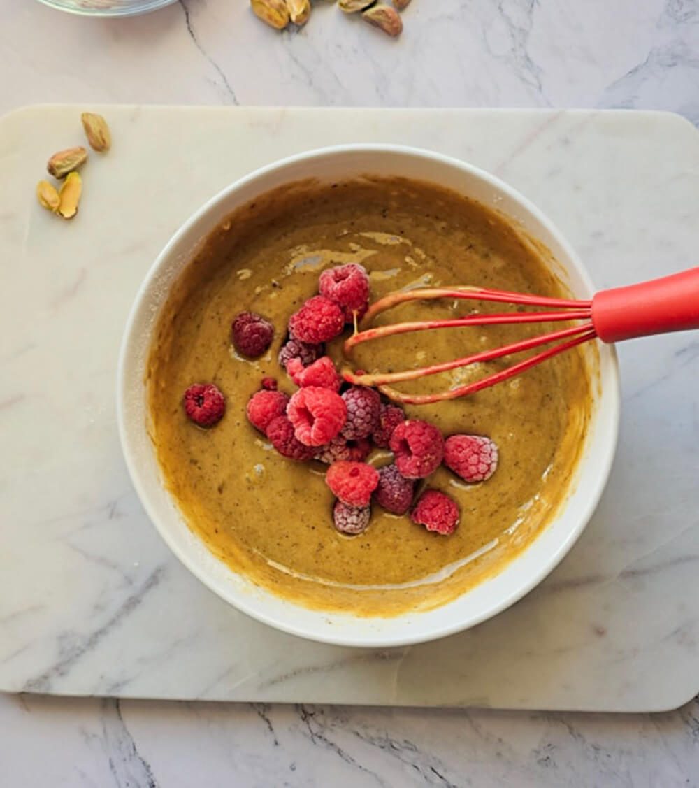 Vue de dessus du bol blanc. La pâte à cake lisse est d'un jaune orangé. Une généreuse poignée de framboises surgelées est déposée au centre. Le fouet rouge est prêt à les incorporer délicatement. Arrière-plan de marbre avec pistaches décoratives.