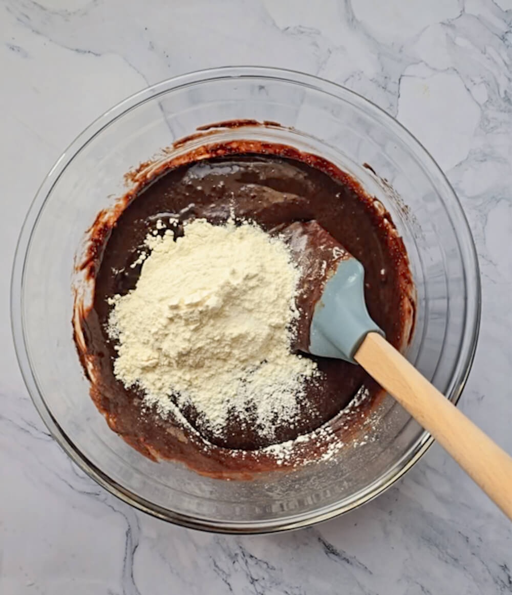 Un bol en verre contenant une préparation pour fondant au chocolat noir mélangée à un monticule de farine blanche, avec une spatule bleu clair à manche en bois prête à incorporer.