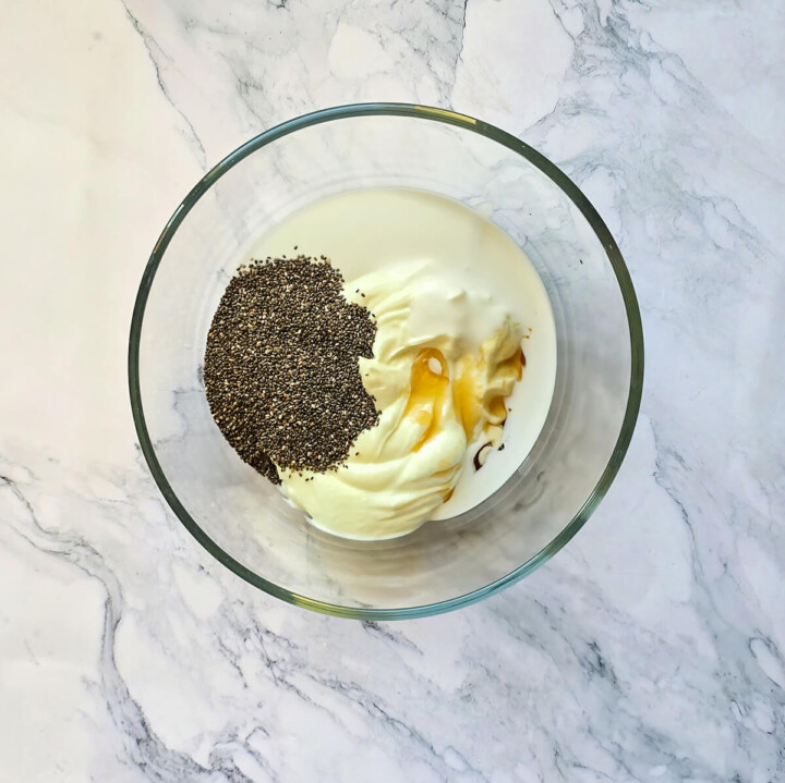 Graines de chia, yogourt grec, lait et sirop d'érable non mélangés dans un grand bol en verre sur un plan de travail en marbre blanc.