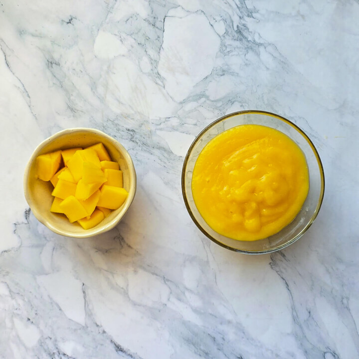 Gros plan d'une mangue entière non pelée et d'un bol de mangue coupée en dés sur un plan de travail en marbre blanc.