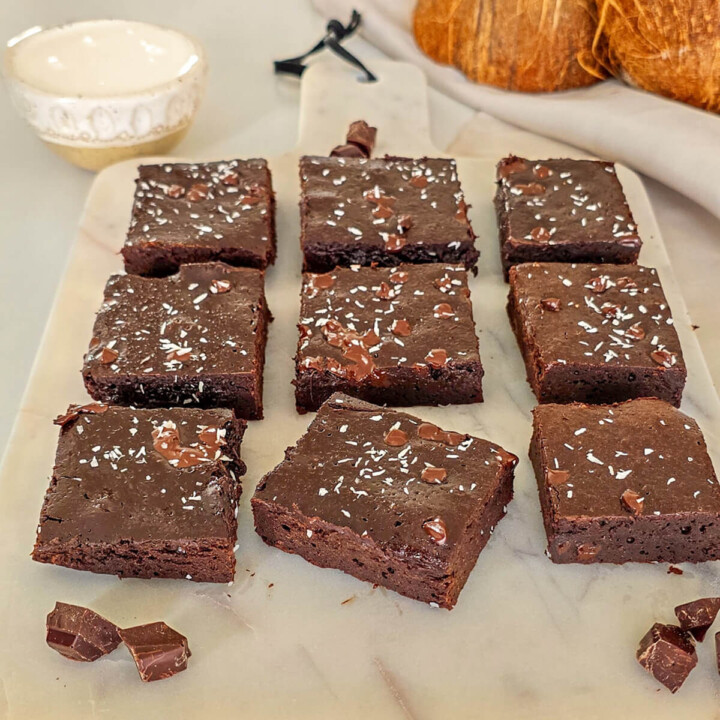 Présentation de neuf parts de fondant chocolat sans beurre crème de coco saupoudrées de noix de coco râpée sur une planche en marbre blanc. La mise en scène inclut une noix de coco entière et un bol de crème pour illustrer cette alternative saine au beurre.