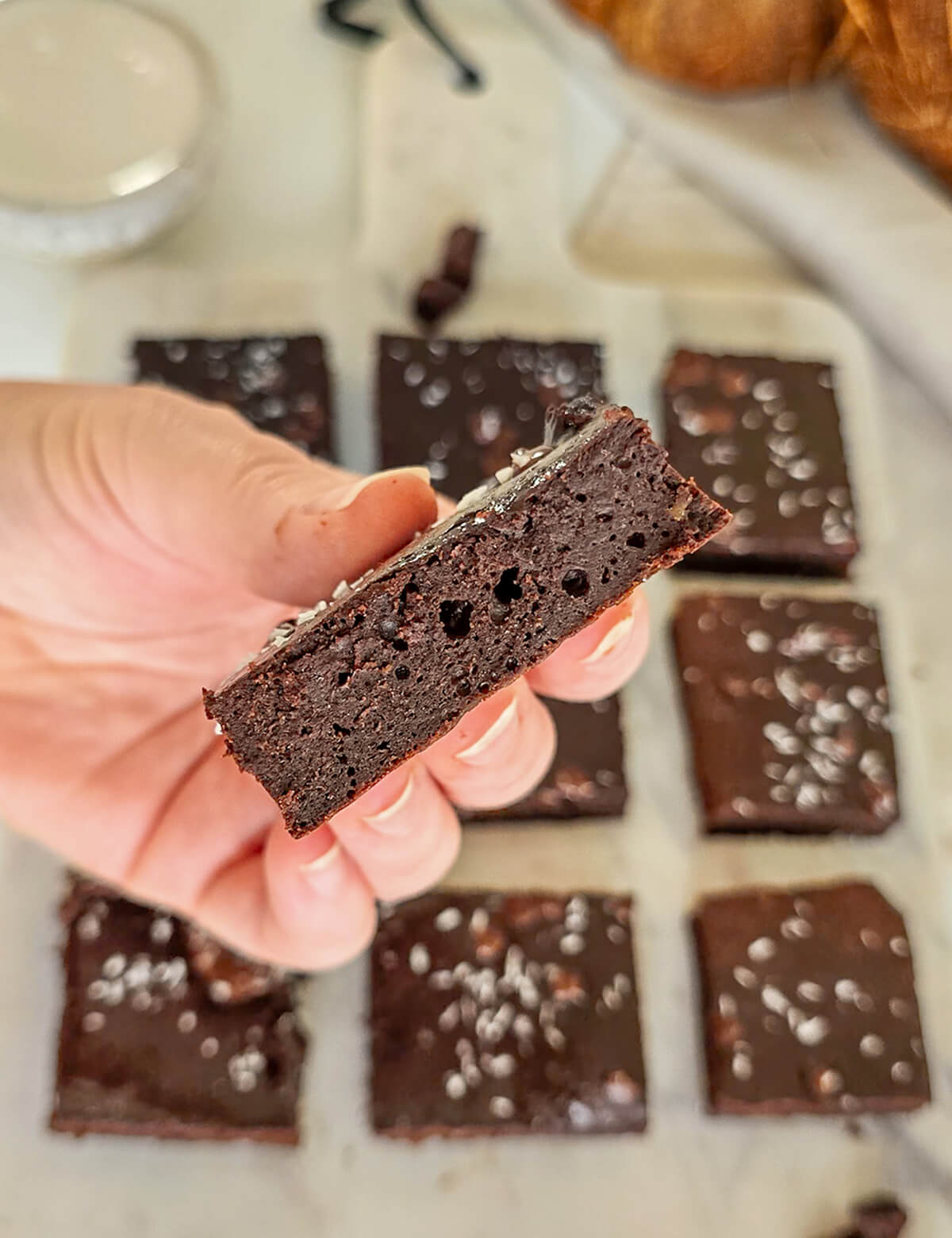 Gros plan sur la texture d'une part de fondant chocolat sans beurre à la crème de coco tenue à la main, révélant un cœur ultra moelleux et fondant. En arrière-plan, les autres carrés de ce gâteau sans lactose sont présentés sur une planche en marbre.