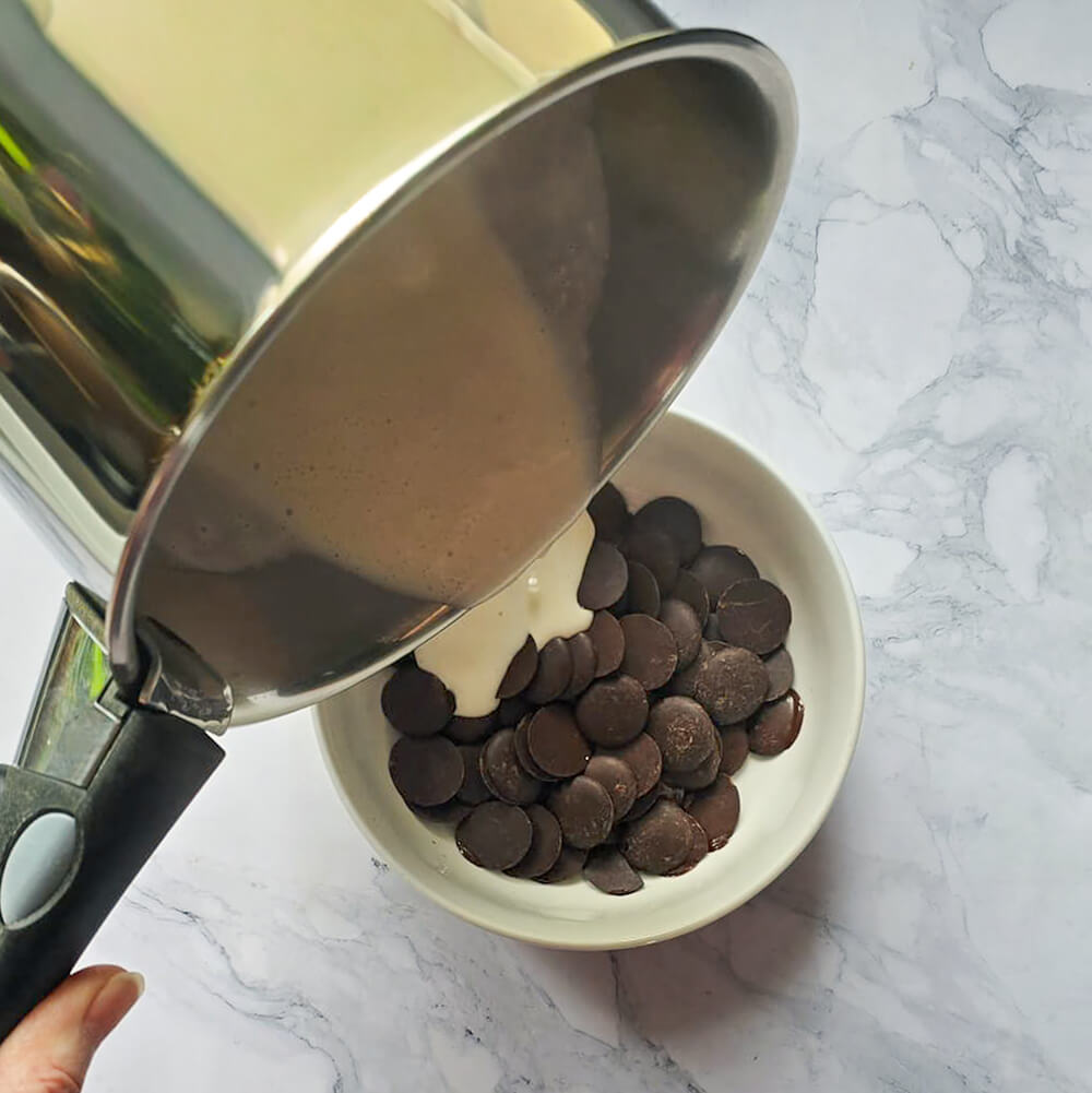 Une main versant de la crème de coco chaude dans un bol rempli de chocolat noir patissier.