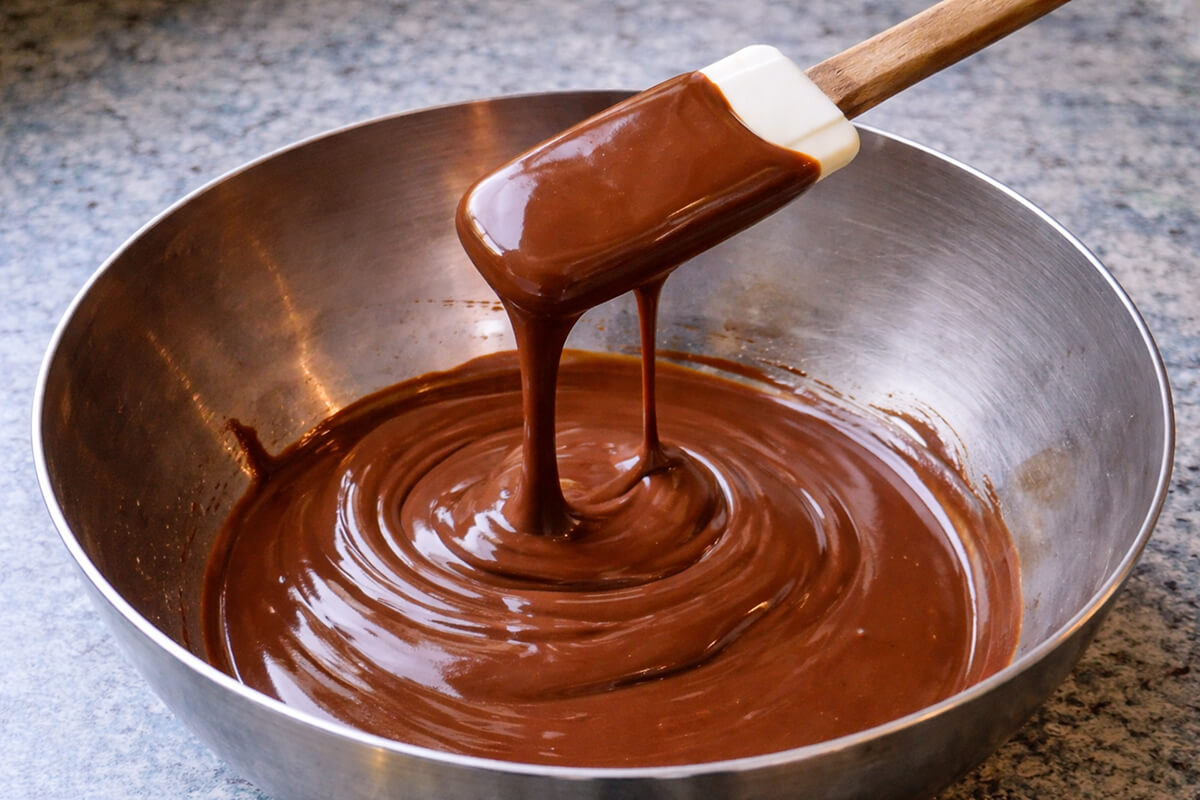 Gros plan d'une ganache au chocolat visiblement trop liquide et trop fluide, s'écoulant en un filet fin d'une spatule en silicone dans un bol en inox, pour illustrer une ganache manquant de prise.