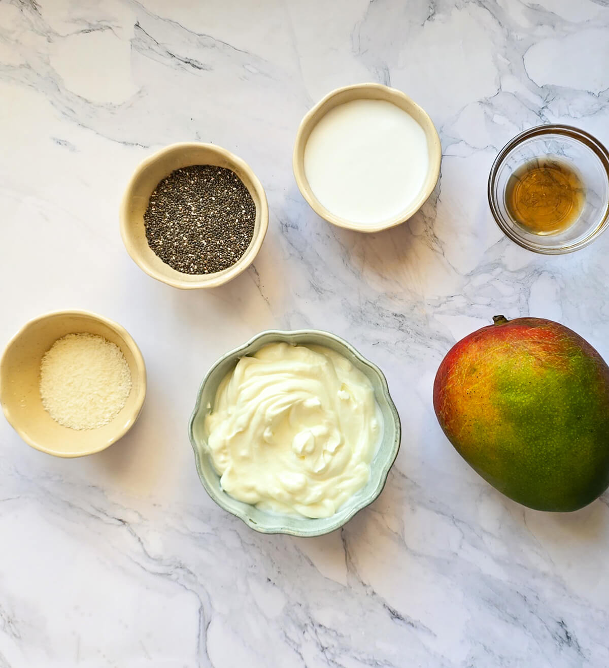 Vue de dessus d'ingrédients disposés dans des bols séparés sur un plan de travail en marbre blanc : graines de chia, lait, sirop d'érable, noix de coco râpée, yogourt grec et une mangue entière.