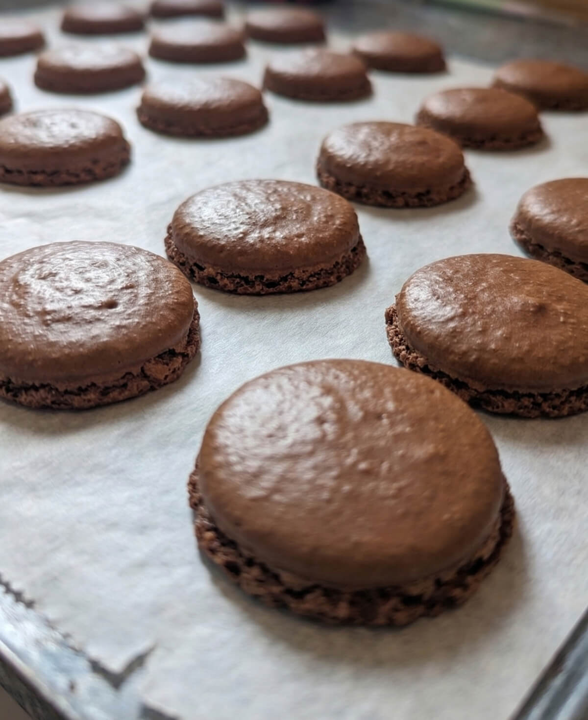 Des coques macarons au chocolat cuits sur une plaque de cuisson, montrant une texture granuleuse, pas assez macaronnés.