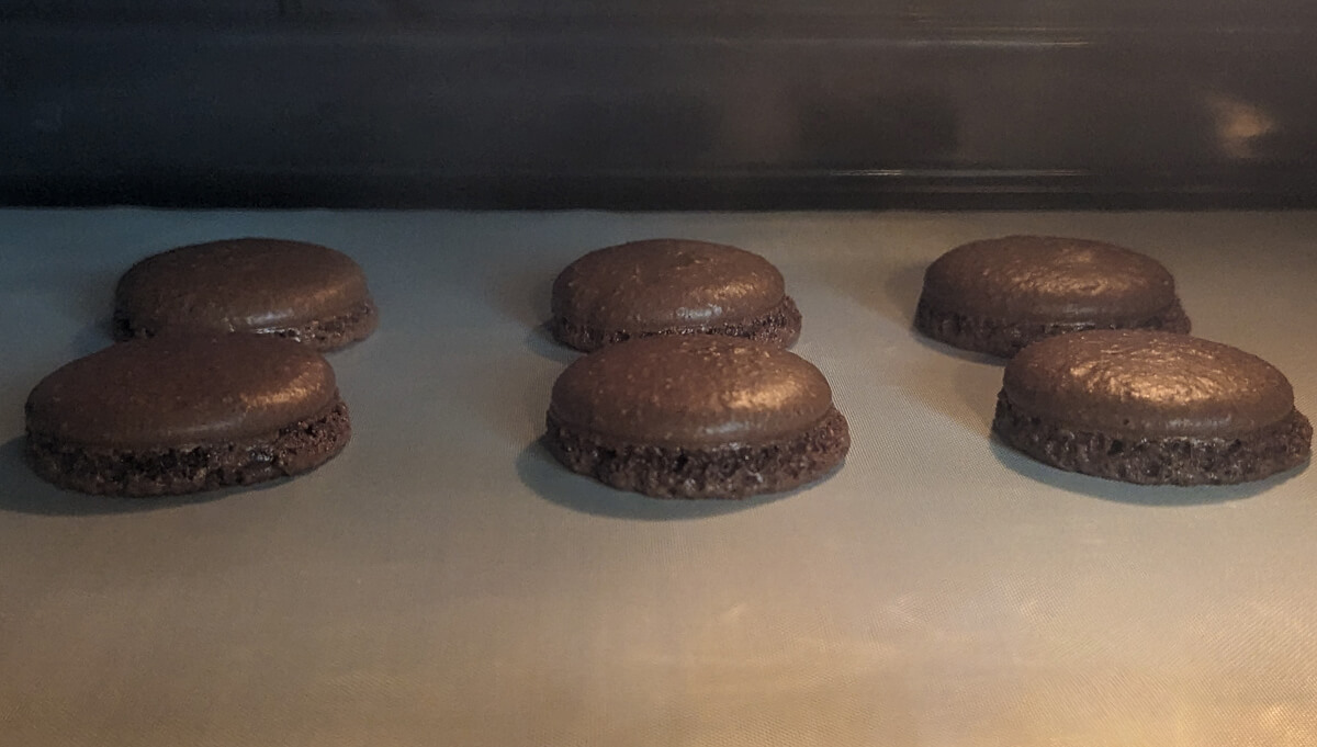 Vue intérieure d'un four montrant une plaque de cuisson avec six coques de macarons au chocolat en pleine cuisson. Les coques, disposées sur un tapis de cuisson en silicone gris, ont une surface lisse et brillante et développent une collerette (le "pied") caractéristique sur leur base. La scène est éclairée par la lumière chaude et tamisée du four, dont le fond sombre est visible.