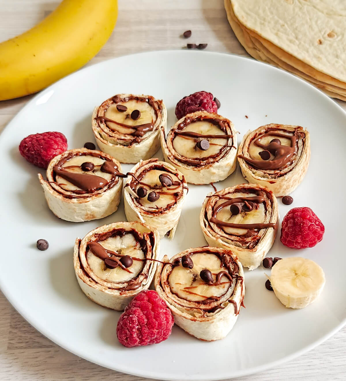 Vue d'ensemble d'une assiette blanche remplie de makis sucrés banane chocolat, entourée d'une banane entière, de pépites de chocolat et d'une pile de tortillas wraps.
