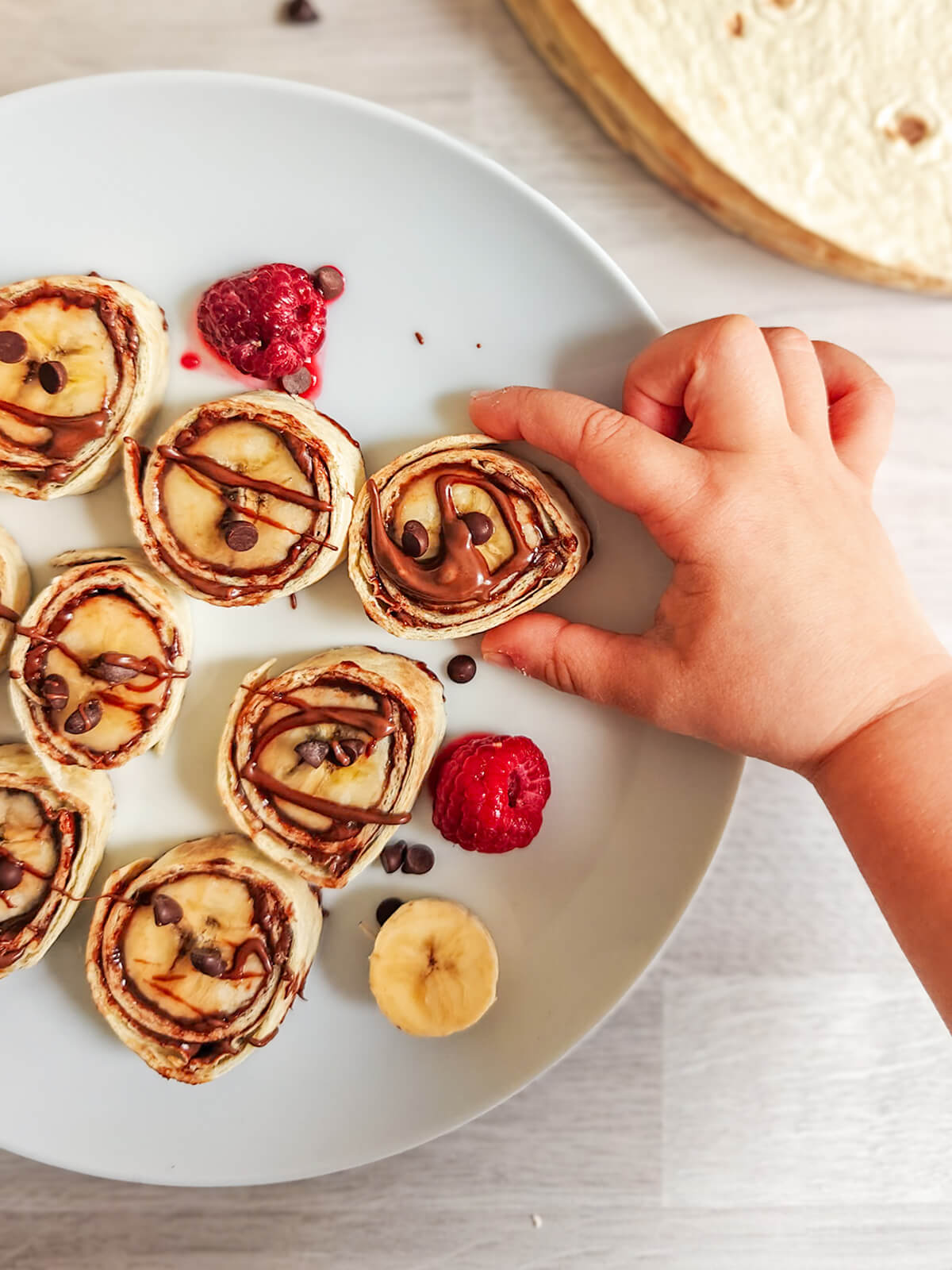 Gros plan sur une assiette de makis Nutella banane avec des framboises fraîches, une main d'enfant attrape un maki gourmand.
