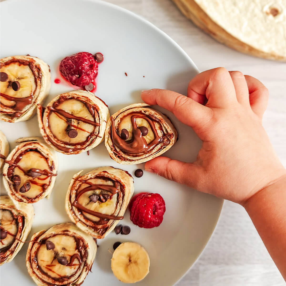 Vue d'ensemble d'une assiette blanche remplie de makis sucrés banane chocolat, entourée d'une banane entière, de pépites de chocolat et d'une pile de tortillas wraps.