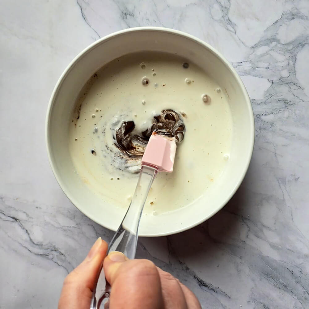 Une main qui mélange une préparation liquide avec une maryse rose, dans un bol blanc.