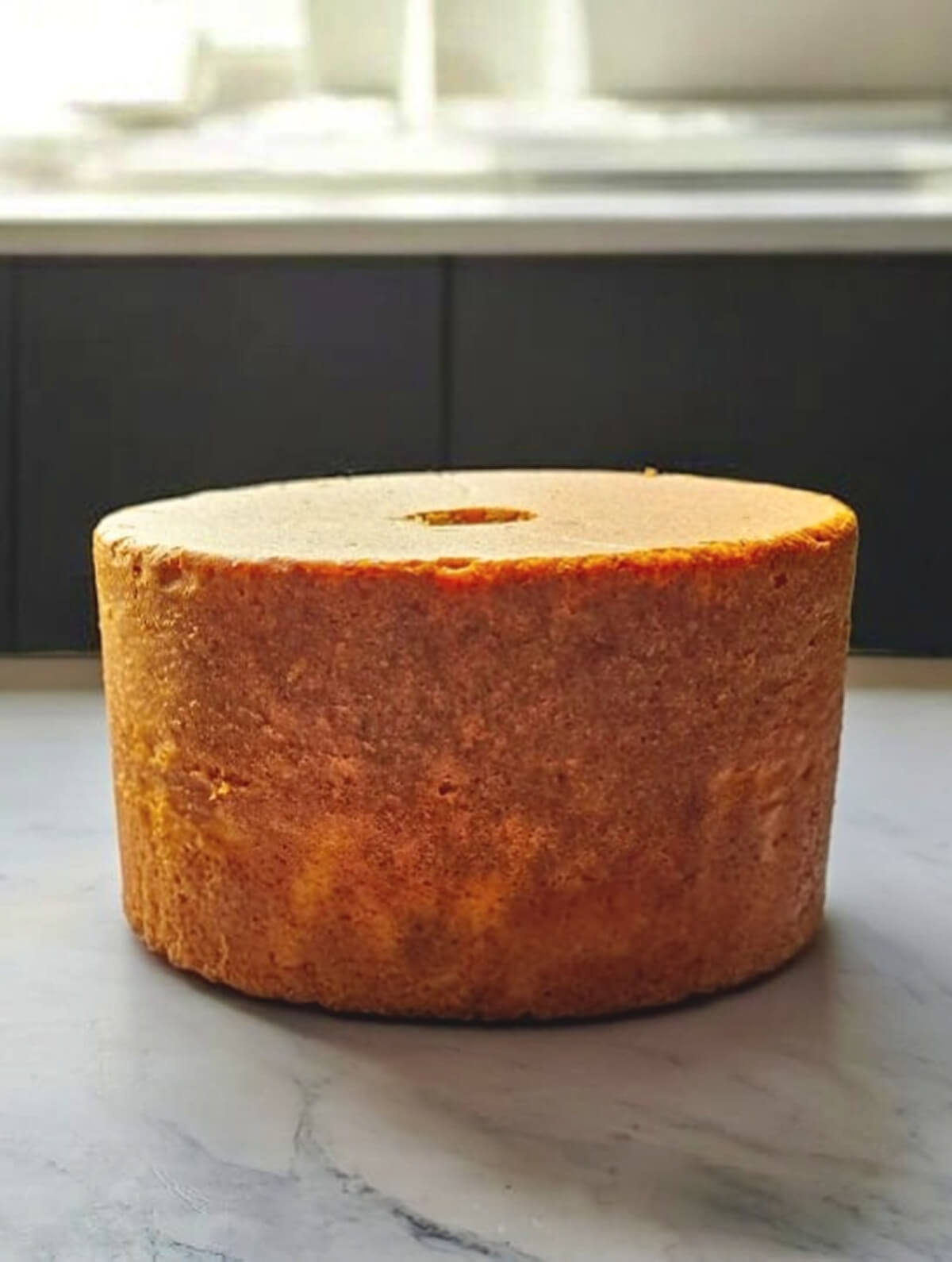 Vue de face d'un grand Molly Cake doré, non glacé, avec un trou central visible, sur un comptoir en marbre dans une cuisine moderne.