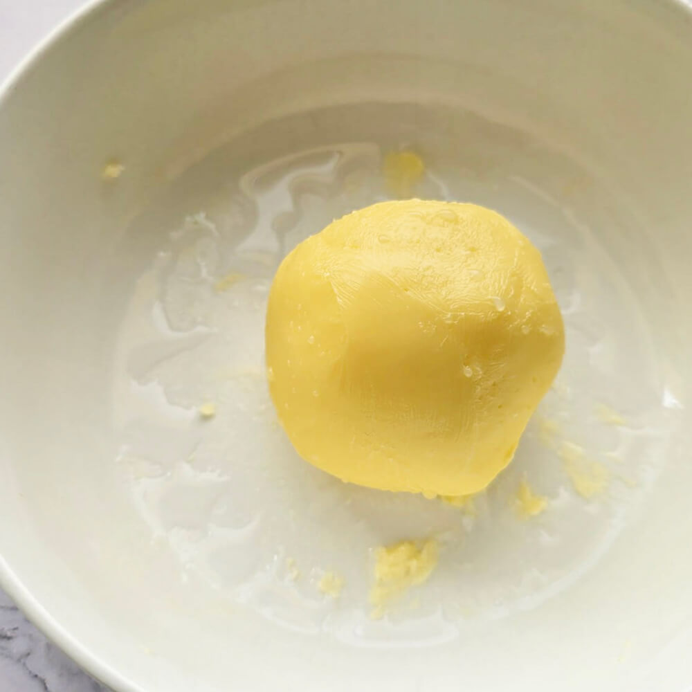 Une vue de dessus d'un bol blanc lisse contenant une belle motte sphérique et homogène de beurre maison jaune pâle. Le beurre a une texture onctueuse et fraîche, et le bol est posé sur un plan de travail en marbre blanc.