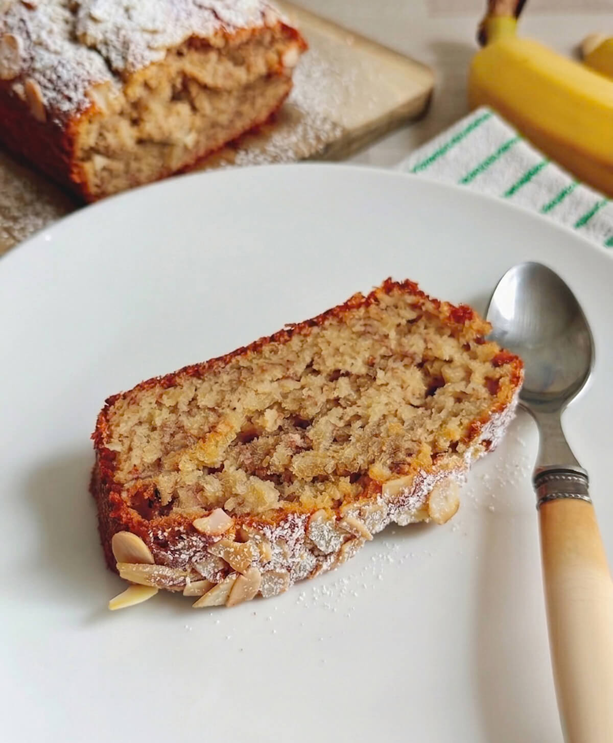 Tranche de banana bread façon frangipane parsemée d'amandes effilées et d'un voile de sucre glace, servie dans une assiette blanche avec une petite cuillère, sur un fond de cuisine lumineux.
