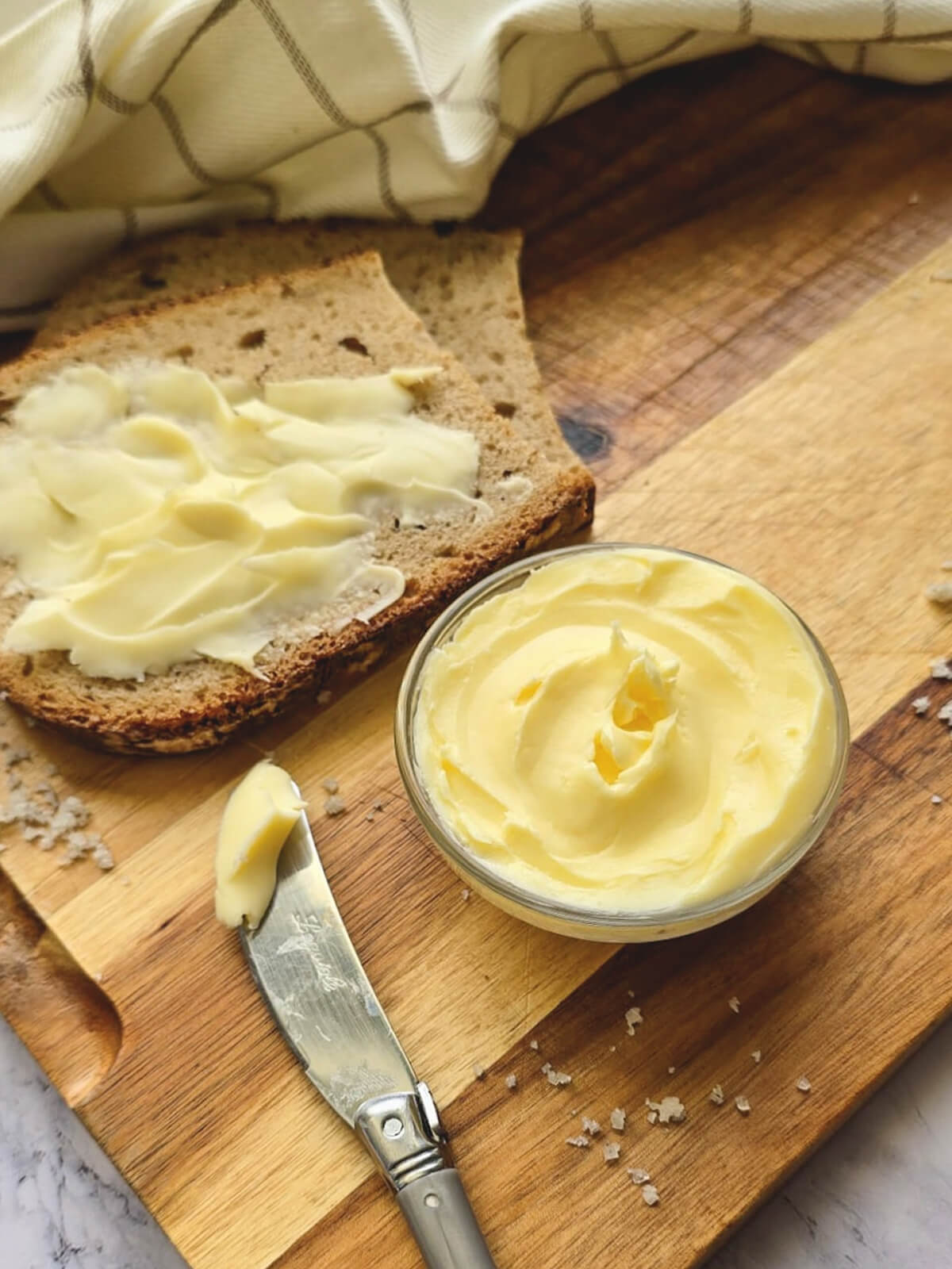 Beurre maison onctueux étalé sur une tranche de pain de campagne, avec un ramequin de beurre et un couteau Laguiole sur une planche en bois.