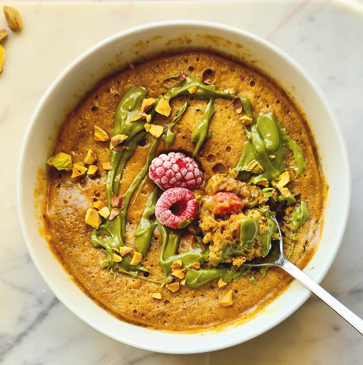 Vue de dessus d'un bowl cake à la pistache et framboise, au moelleux apparent, servi dans un bol blanc sur un marbre clair. Il est généreusement nappé d'une crème de pistache vert vif et parsemé d'éclats de pistaches grillées. Au centre, deux framboises surgelées sont entourées de baies cuites.