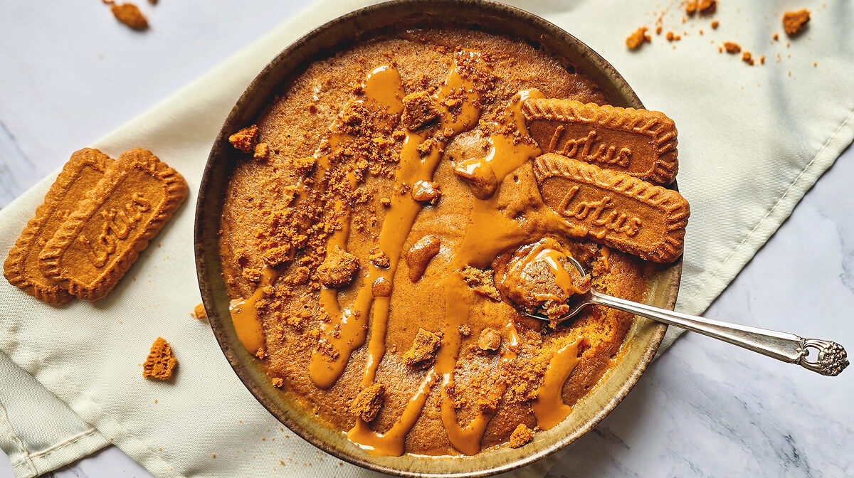 Vue de dessus d'un bowlcake au spéculoos bien doré, décoré d'un filet de pâte à tartiner onctueuse et de morceaux de biscuits. Une cuillère en argent prélève une bouchée gourmande au centre du bol.